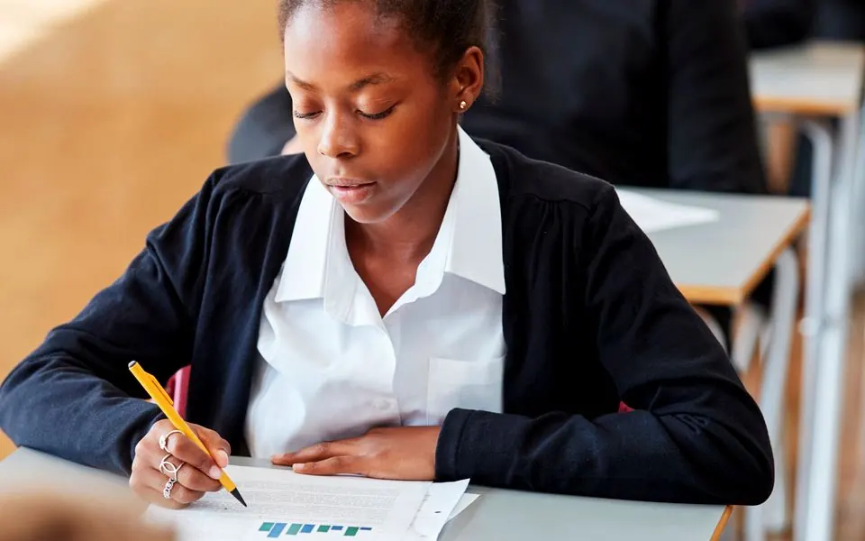 A school learner writing at a desk, other learners working behind her.