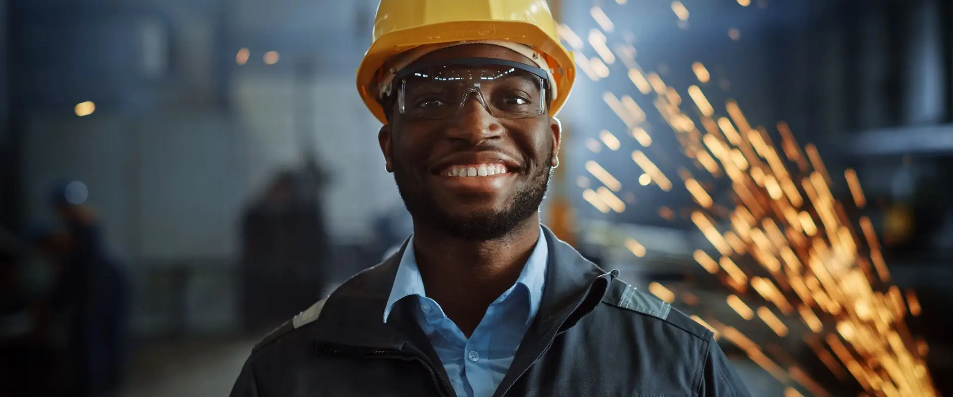 A worker in a hard hat and safety glasses, sparks flying from a cut behind them.